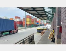 Außenbereich eines Industrie- oder Logistikgebäudes mit Backsteinfassade, überdachtem Gehweg und Blick auf eine Straße. Im Hintergrund sind zahlreiche bunte Frachtcontainer gestapelt, daneben ein LKW mit Containeraufbau. Auf dem Gehweg befinden sich Mülltonnen und Paletten.