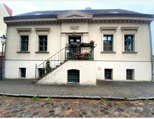 Zweistöckiges historisches Gebäude mit heller Fassade, symmetrisch angeordneten Fenstern und schmuckvollem Gesims. Eine Treppe führt zu einem kleinen Balkon mit Blumen, darunter ein unterer Bereich mit kleinen Fenstern und einem Gitter; Kopfsteinpflaster und eine Laterne rahmen den Eingangsbereich.
