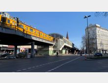 Gelbes U-Bahn-Fahrzeug auf einer erhöhten Brücke der Berliner Hochbahn, daneben ein historischer U-Bahnhof mit großen Fenstern und gewölbter Dachkonstruktion; im Hintergrund ist eine Kirchturmspitze und ein Altbau zu sehen. Straßenverkehr und Fußgänger prägen das urbane Umfeld.