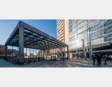 Außenansicht des Bahnhofs Potsdamer Platz in Berlin – eine moderne Glas-Stahl-Konstruktion mit überdachtem Eingang, Rolltreppen und Fahrradständern. Im Hintergrund hohe Bürogebäude mit Glasfassaden sowie Passanten auf dem Platz.