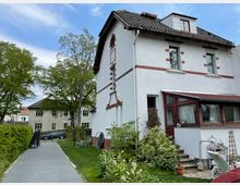 Zweistöckiges Wohnhaus mit weißer Fassade und roten Backsteindetails, ergänzt durch einen verglasten Wintergarten. Neben dem Haus eine gepflasterte Einfahrt, die von Hecken und Bäumen gesäumt ist, sowie ein kleiner Garten mit Spielgeräten.