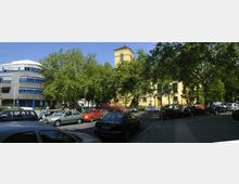 Straßenszene mit parkenden Autos und Fußgängern in einer urbanen Umgebung, umgeben von Bäumen. Im Hintergrund links ein modernes Gebäude mit blauer und weißer Fassade, rechts eine gelbe Kirche mit Turm, teilweise verdeckt durch das Laub der Bäume.