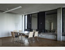 Moderner Besprechungsraum mit einem rechteckigen Tisch und fünf Holzstühlen auf einem Boden aus Parkett. Eine Wand mit großen Fenstern und schwarzen Lamellen sowie eine weiße Wand mit minimalistischer Dekoration rahmen den Raum, der von schlichten Deckenleuchten beleuchtet wird.