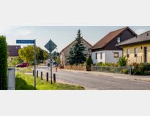 Straßenszene in einem Wohngebiet mit Einfamilienhäusern, Vorgärten und einem Straßenschild mit der Aufschrift „Am Schützenpark“. Die Straße ist asphaltiert, flankiert von Bäumen und Zäunen, und im Hintergrund stehen geparkte Autos.
