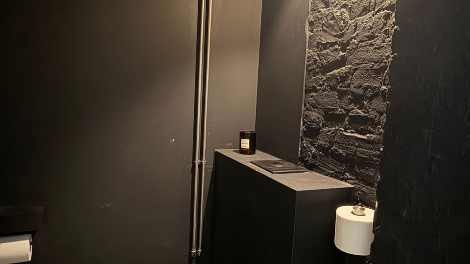 Kleines, dunkel gestaltetes Gäste-WC mit schwarzer Wandtoilette, Betonboden und sichtbaren Metallrohren an der Wand. Eine freigelegte Ziegelwand mit Wandleuchte, schmalem Ablagekasten sowie Toilettenpapierhalter und WC-Bürste ergänzen den Raum.