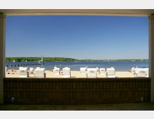 Blick auf einen Sandstrand mit nummerierten Strandkörben vor einem See. Im Hintergrund sind ein langer Steg, Segelboote und bewaldete Ufer zu sehen, unter klarem, blauem Himmel.