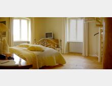 Helles Schlafzimmer mit einem Eisenbett und gelber Tagesdecke, zwei großen Fenstern mit Vorhängen, einem hölzernen Schreibtisch, einem kleinen Fernseher auf einer Kommode und einer Wendeltreppe in der Ecke. Der Boden ist aus hellem Holz.