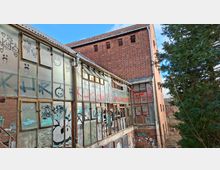 Verlassener Industrie- oder Lagerbau aus rotem Backstein mit einem langen Anbau aus Glas- und Metallrahmen, dessen Scheiben teils beschädigt und mit Graffiti bedeckt sind. Im Hintergrund ragt ein hoher Backsteinturm mit kleinen Fensteröffnungen auf, daneben stehen dichte Bäume am Rand des Geländes.