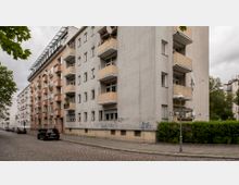 Mehrstöckiges Wohnhaus in einer städtischen Straße, gebaut aus grauem Putz mit mehreren Balkonen. Im Vordergrund ist eine gepflasterte Straße mit parkenden Autos zu sehen, während Straßenschilder und eine Laterne die Ecke markieren; umgeben von Bäumen und Sträuchern.