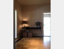 Ein Eingangsbereich mit hellbeigem Fliesenboden, einem schwarzen Konsolentisch an der Wand, darauf eine Tischlampe und eine Pflanze. Im Hintergrund ist eine geöffnete Tür mit Blick auf einen hellen, grünlichen Außenbereich.