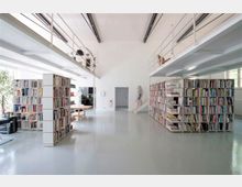 Moderner, heller Bibliotheksraum mit hohen Decken, zwei großen Bücherregalen in der Mitte und einer umlaufenden Galerie mit Geländer. Tageslicht fällt durch große Fenster ein, und die Einrichtung umfasst auch Sitzbereiche und einen Tisch im Hintergrund.