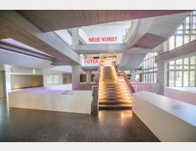 Innenansicht eines modernen Museums mit Sichtbetonarchitektur, einer zentralen beleuchteten Treppe und großen Fenstern, die viel natürliches Licht hereinlassen. Beschilderungen mit den Worten „NEUE KUNST“ und „FOYER“ sind sichtbar.