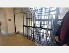 Blick auf ein Treppenhaus mit Metallgittern, die den Treppenbereich absperren. Im Hintergrund ist natürliche Beleuchtung durch große Fenster sichtbar, der Boden besteht aus hellem Laminat.