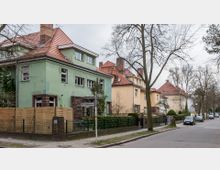 Straßenszene mit mehreren zweigeschossigen Wohnhäusern im Stil der 1920er bis 1930er Jahre, darunter ein grünes Haus im Vordergrund mit rotem Ziegeldach und eingezäuntem Vorgarten. Die Straße ist von Bäumen gesäumt, und am Gehweg stehen geparkte Autos.