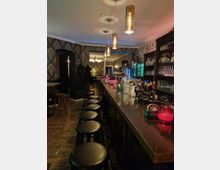 Innenansicht einer stilvollen Bar mit langer, dunkler Theke, davor schwarzen Barhockern. Im Hintergrund sind eine Tapete mit floralem Muster, beleuchtete Regale mit Flaschen, hängende Lampen sowie ein Bereich mit Tischen und Stühlen zu sehen.