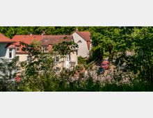 Mehrfamilienhäuser mit roten Ziegeldächern, umgeben von dichter Vegetation und Bäumen; im Vordergrund Sträucher, im Hintergrund ein kleiner Parkplatz mit einem roten Fahrzeug.