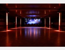 Ein leerer Veranstaltungssaal mit einem Holzboden, seitlichen Holzsäulen und einer erhöhten Bühne im Hintergrund. Der Raum ist dunkel beleuchtet, mit bunten Scheinwerfern, die auf die Bühne gerichtet sind.