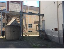 Ein industrielles Außengelände mit einem massiven Betonbau in Kegelform, umgeben von Stahlträgern und Gebäudewänden mit sichtbaren Ziegel- und Putzflächen. Eine Metalltreppe führt zu einer Tür in einer weiß gestrichenen Wand mit sichtbaren Gebrauchsspuren.