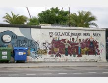 Ein Abschnitt der Berliner Mauer mit bemalter Kunst, die Figuren zeigt, die eine rote Ziegelmauer bauen, begleitet vom Schriftzug „Es gilt viele Mauern abzubauen“. Im Vordergrund stehen Müllcontainer, und im Hintergrund sind Palmen und Bäume sichtbar.