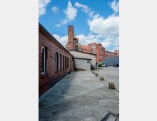 Ein Industriehof mit Backsteingebäuden, einem Schornstein und einer gepflasterten Fläche. Im Hintergrund sind mehrere Gebäude mit teils roten Dächern sowie parkende Autos zu sehen, der Himmel ist blau mit einigen Wolken.