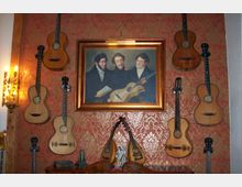 Wand mit roter, gemusterter Tapete, dekoriert mit antiken Gitarren und zwei Mandolinen, sowie einem in Gold gerahmten Gemälde dreier Männer mit einer Gitarre. Links ist eine Wandleuchte im klassischen Stil zu sehen, darunter stehen kleine dekorative Gegenstände auf einem Holztisch.