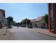Straßenszene in einer kleinen Stadt mit Kopfsteinpflaster, flankiert von Häusern mit roten Ziegeldächern und kleinen Geschäften. Links und rechts der Straße stehen Bäume, und einige Autos sind geparkt; in der Mitte fahren zwei Fahrradfahrer.