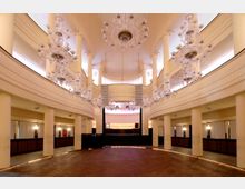 Innenansicht eines großen, eleganten Veranstaltungsraums mit hölzernem Parkettboden, hohen Säulen und einer Galerie auf beiden Seiten. Mehrere gläserne Kronleuchter hängen von der gewölbten Decke, während eine erhöhte Bühne mit Beleuchtung im Hintergrund sichtbar ist.