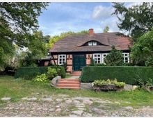 Ein Fachwerkhaus mit Backsteinfassade und dunklem Giebeldach, umgeben von einer Hecke und Bäumen. Vor dem Haus führt eine kurze Treppe mit roten Ziegelstufen zum grünen Eingang, während der Garten von Steinen und Pflanzen geprägt ist.
