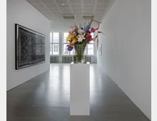 Ein moderner Ausstellungsraum mit glatten Betonböden und weißen Wänden, in dem ein farbenfrohes Blumenarrangement auf einem weißen Podest im Vordergrund steht. An der linken Wand hängt ein großformatiges Bild mit schwarzem, ornamentalem Muster, und im Hintergrund befinden sich große Fenster, die Tageslicht in den Raum lassen.