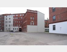 Backsteinfassaden eines mehrstöckigen Gebäudekomplexes mit weißen und roten Elementen, flankierend auf einem offenen, asphaltierten Hof. Im Hintergrund ein Durchgangsbogen mit einem kleinen Schild und einigen Fenstern mit Sprossenglas.