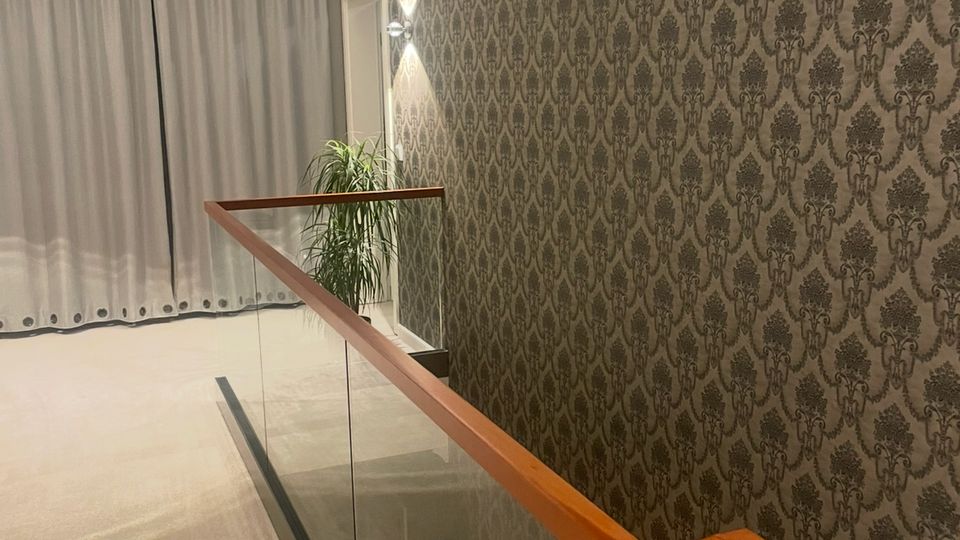 Ein modern gestalteter Flur mit Teppichboden, einer gläsernen Treppenbrüstung mit Holzhandlauf und dekorativer Tapete mit floralen Mustern an der Wand. Im Hintergrund sind geschlossene, weiße Vorhänge und eine Zimmerpflanze sichtbar.
