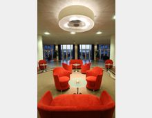 Lounge-Bereich mit roten Polstersesseln, runden Glastischen und großem, zentralem Deckenleuchter. Im Hintergrund sind große Fensterfronten und Topfpflanzen sichtbar.