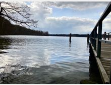 Ein Holzsteg mit Metallgeländer ragt in einen ruhigen See, umgeben von Wald am Ufer, unter einem Himmel mit dichten Wolken und vereinzelten Lichtungen.