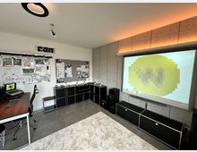 Moderner Arbeitsraum mit einem Eckschreibtisch, schwarzen Möbeln und einer Betonwand. Ein Projektor zeigt ein Bild auf eine Leinwand an der rechten Wand, und die Decke hat eine minimalistische Rundlampe sowie eine dezente Hintergrundbeleuchtung.