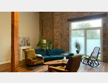 Wohnzimmer mit Backsteinwand, einem großen Fenster mit Blick nach draußen und einem Holzboden. Die Einrichtung umfasst eine grüne Couch, zwei gelbe Sessel, einen Schaukelstuhl, einen Holztisch und mehrere Zimmerpflanzen.