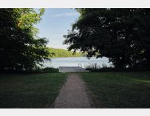 Ein schmaler Weg führt durch eine Wiese zu einem kleinen Holzsteg mit Geländer, der in einen von Bäumen umgebenen See ragt. Der Steg liegt zwischen hohen Büschen und Schatten spendenden Bäumen, mit Blick auf die gegenüberliegende bewaldete Uferseite.