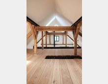 Dachgeschoss eines Gebäudes mit sichtbaren Holzbalken und hellem Holzfußboden. Das spitze Dach hat weiße Wände und ein kleines rechteckiges Fenster am Ende des Raumes.