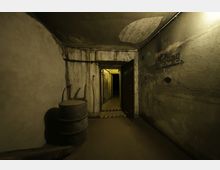 Ein unterirdischer Betonraum mit abgenutzten Wänden und einer offenen Tür, die in einen erleuchteten Gang führt. Im Vordergrund stehen zwei Metallfässer, an der rechten Wand ist das Wort "Eingang" mit einem Pfeil erkennbar.