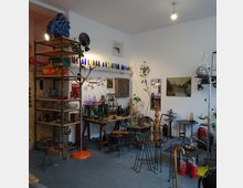 Ein kleiner Laden oder Atelier mit einer vielseitigen Sammlung von dekorativen Gegenständen, vor allem Glaswaren wie Vasen und Flaschen in verschiedenen Farben. Der Raum ist mit Regalen, Tischen und Stühlen ausgestattet, die unregelmäßig angeordnet sind, und die Wände sind mit Kunstwerken und Dekorationen geschmückt.