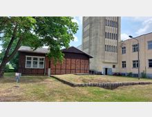 Fachwerkgebäude mit roten Ziegeln und dunklem Holz, daneben ein grauer, hoher Betonturm mit schmalen Fenstern; im Hintergrund ein beiger Gebäudeteil mit mehreren rechteckigen Fenstern. Die Umgebung ist von einer Wiese und einzelnen Bäumen geprägt.