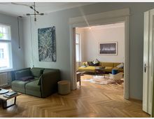 Wohnzimmer mit modernem Design, bestehend aus einem grünen Sofa, einem gelben Ecksofa und einem hellen Holzboden im Fischgrätenmuster. Große Fenster lassen viel Tageslicht herein, und an den Wänden hängen gerahmte Bilder.