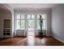 Heller, leerer Wohnraum mit hohen Decken, Stuckleisten und großem dreiteiligem Fenster mit Balkontür zur Terrasse. Links ein Einbauschrank in einer Nische, unter den Fenstern Heizkörperverkleidungen und rechts ein großer Kaktus im Topf auf Holzfußboden.