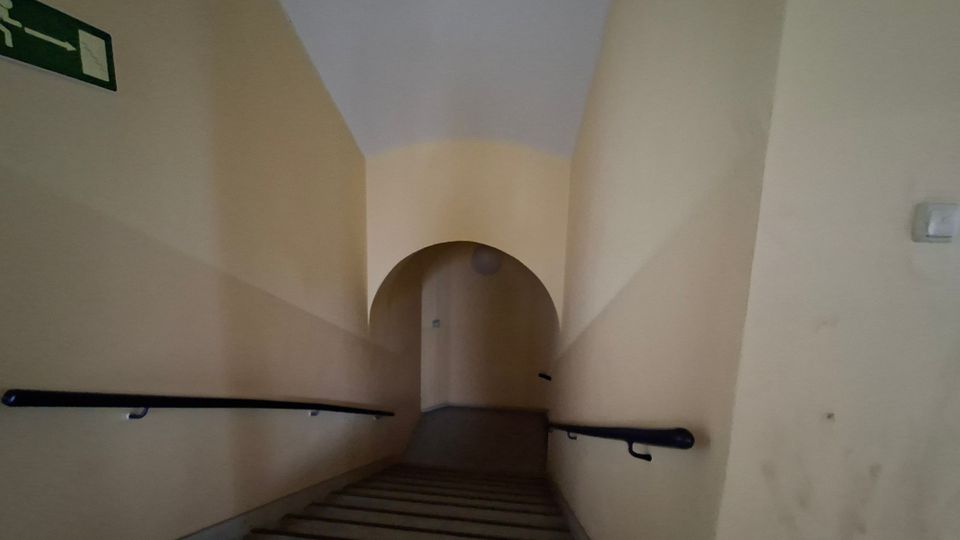 Schmales Treppenhaus mit hellbeigen Wänden und Handläufen an beiden Seiten, das nach oben zu einem rundbogigen Durchgang führt. Die Decke wölbt sich leicht, und am Ende des Aufgangs ist eine Tür im Halbrund sichtbar.