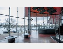 Innenraum eines modernen Gebäudes mit großer Glasfassade, die den Blick auf einen Fluss mit Booten und einer Baumlandschaft freigibt; im Vordergrund befinden sich runde Sitzmöbel und eine geschwungene Bar mit Flaschenregalen, rechts eine Wendeltreppe.