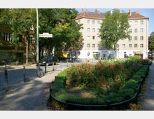 Ein kleiner Stadtplatz mit einem bepflanzten Blumenbeet, umgeben von niedrigen Pollern und einem Pflasterweg. Im Hintergrund befinden sich ein mehrstöckiges Wohngebäude mit heller Fassade, ein Straßenschild und vereinzelt geparkte Fahrzeuge sowie Bäume, die Schatten spenden.