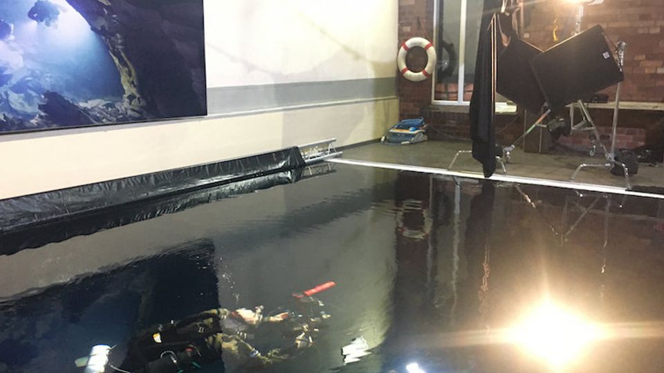 Indoor diving training pool with dark water, bordered by black plastic sheeting and surrounded by brick and white walls. Studio lighting equipment and a wall-mounted life ring are visible around the pool’s edge.