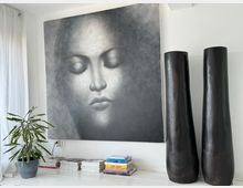 Wohnraum mit minimalistischem Dekor; an einer weißen Wand hängt ein großformatiges monochromes Gemälde eines geschlossenen Gesichts. Darunter steht ein weißes Regal mit Büchern, Zeitschriften und einer Zimmerpflanze. Rechts daneben stehen zwei hohe, dunkle Holzvasen.