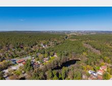 Luftaufnahme einer weitläufigen Waldlandschaft mit verstreuten Wohnhäusern und einer Bahntrasse am Rand einer kleinen Siedlung. Im Vordergrund ist ein dunkler, ovaler Teich von Bäumen umgeben, im Hintergrund reicht der Blick bis zu einer entfernten Stadt am Horizont.