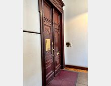 Holztür eines historischen Gebäudes mit aufwendigen Verzierungen und Schlitzfenster-Elementen im oberen Bereich. Ein Metallschild mit der Aufschrift „Lubberger Lehment“ ist an der linken Tür angebracht, der Eingangsbereich hat einen Teppich und einen Parkettboden.