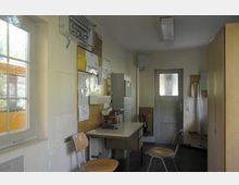 Ein einfacher Büroraum mit einem Tisch, zwei Holzstühlen und einer Pinnwand, die mit Dokumenten und Notizen bestückt ist. Ein Fenster auf der linken Seite lässt Tageslicht herein, und eine Tür mit Milchglas führt aus dem Raum, ergänzt durch eine Wand mit Schaltschränken und einer Feuerlöscheinheit.